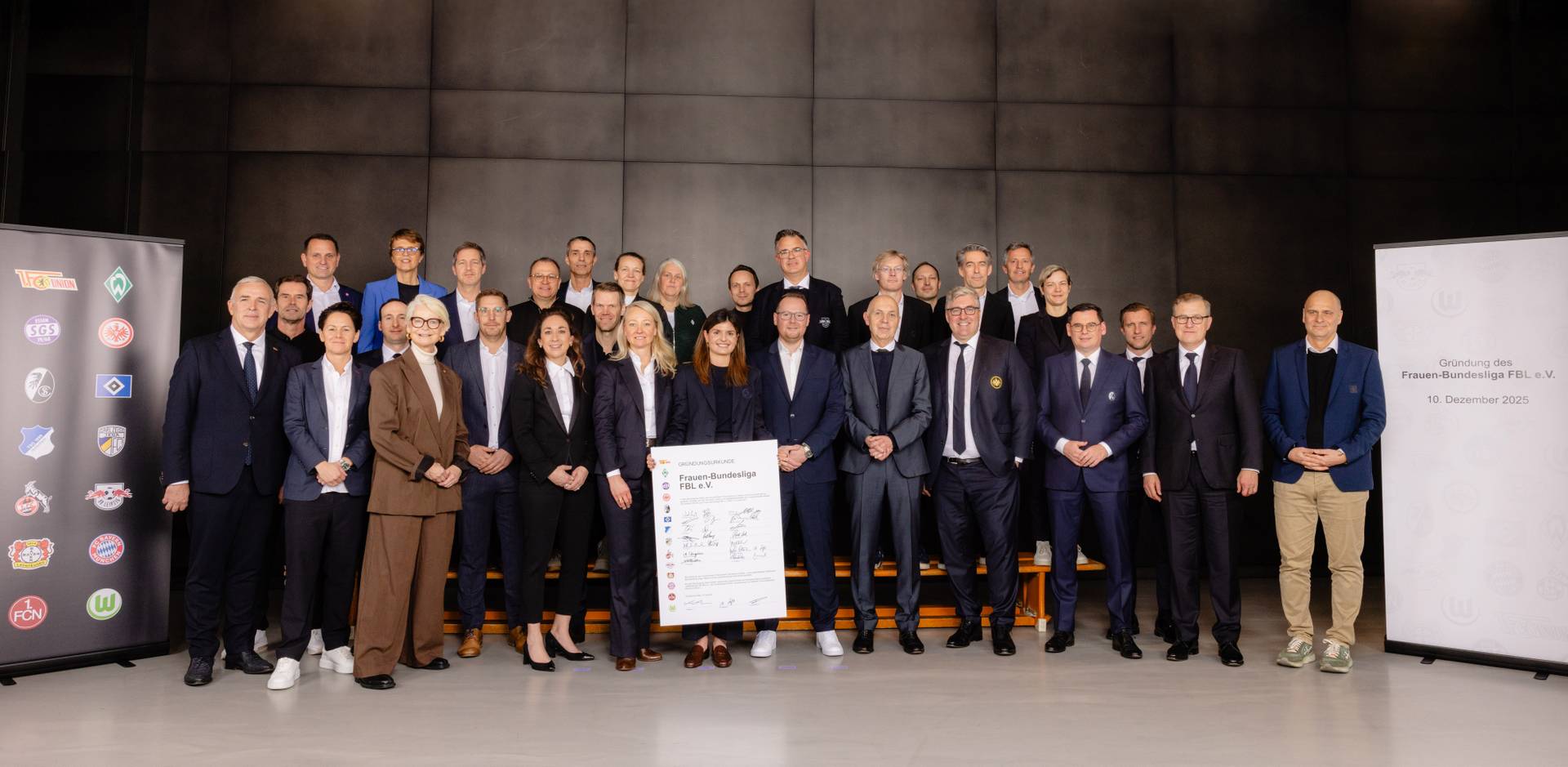 Etwa 30 Geschäftsleute in Anzügen und Kleidern posierten in Reihen im Innenbereich, in der Mitte hielten sie ein großes Plakat, flankiert von Werbebannern.