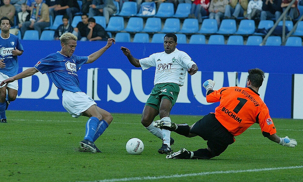 null Ailton mit dem Ball am Fuß auf dem Weg zum Tor. Links von ihm ist ein Bochumer Verteidiger, rechts wirft sich Bochums Torwarm im orangen Trikot auf den Rasen.