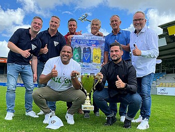 Die Veranstalter halten das Plakat und den Pokal. Ailton und Dieter Burdenski zeigen den Daumen nach oben.