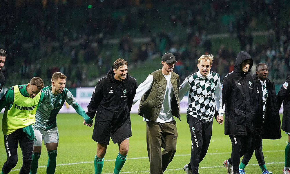 Die Spieler stehen Hand in Hand auf dem Rasen und laufen zur Laola-Welle an.