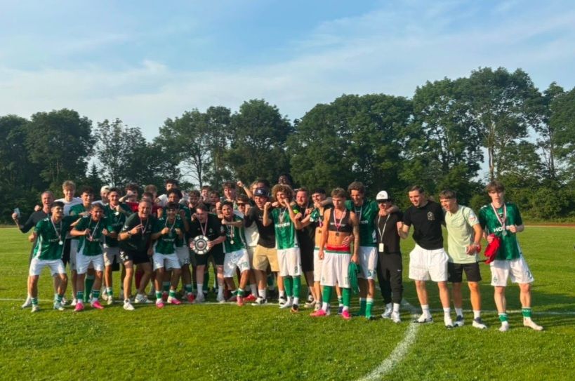 Eine jubelnde Fußballmannschaft posiert gemeinsam auf einem Spielfeld, mit Medaillen und grünen Trikots bekleidet, und feiert ihren Sieg vor dem Hintergrund von Bäumen.