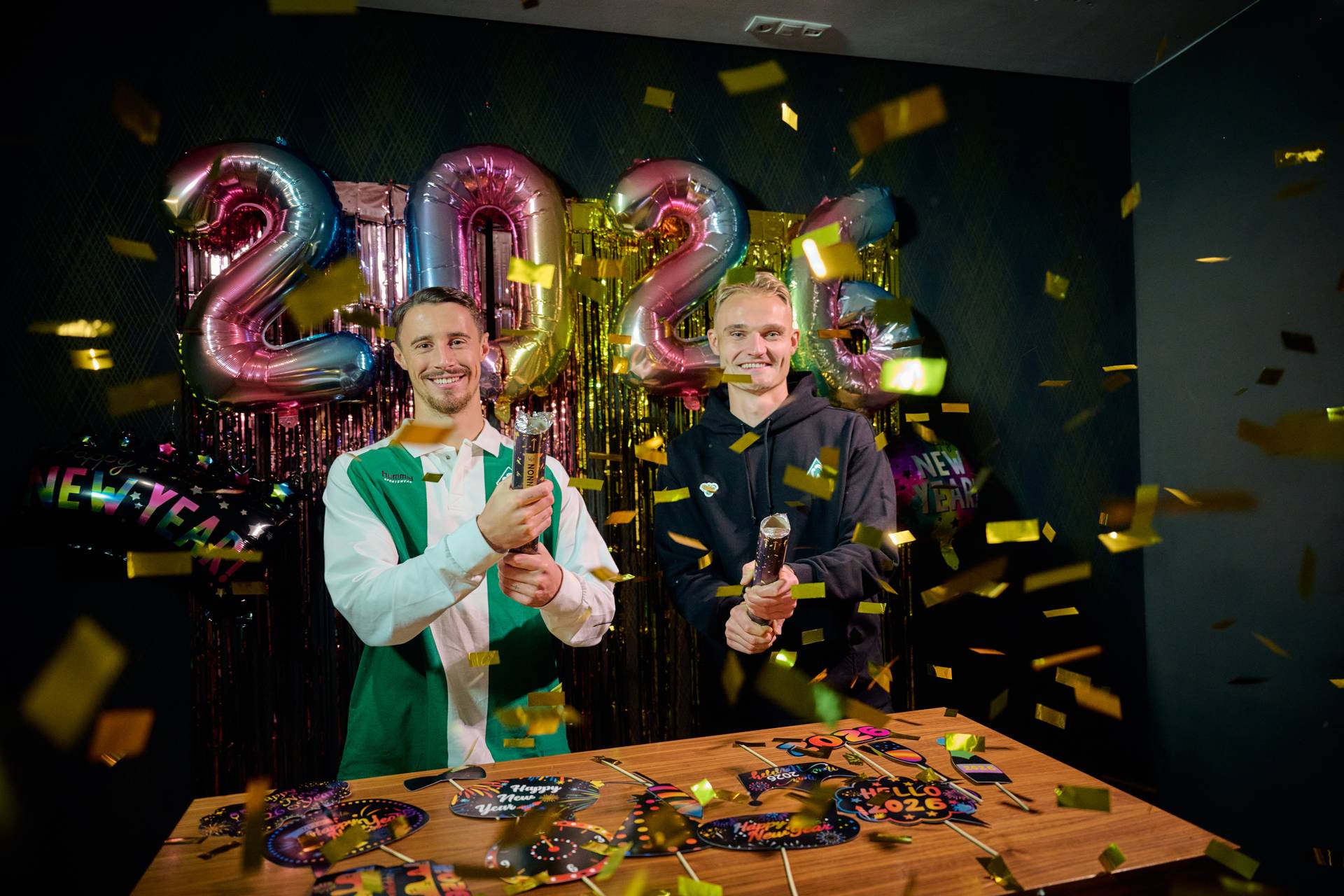 Marco Friedl und Amos Pieper mit einer Konfetti-Kanone, im Hintergrund Luftballons in 2026 Form.