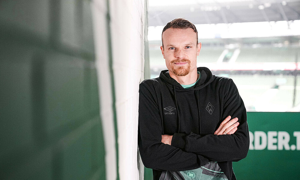 Christian Groß lehnt an einer Wand, im Hintergrund das wohninvest WESERSTADION. 