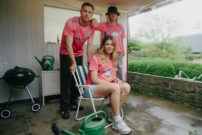 Drei Personen in rosa Trikots auf einer Terrasse, eine sitzt und zwei stehen, im Hintergrund ein Garten und ein Stadion.