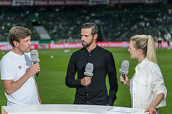 null Martin Harnik steht mit einem Sport1 Mikrofon in der Hand an einem Tisch im wohninvest WESERSTADION. Er steht zwischen Clemens Fritz und Moderatorin Ruth Hofmann.