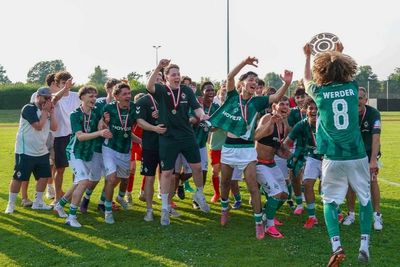 Eine fröhliche Fußballmannschaft in grünen Trikots feiert auf dem Spielfeld; ein Spieler hält einen Pokal in die Höhe, alle tragen Medaillen.