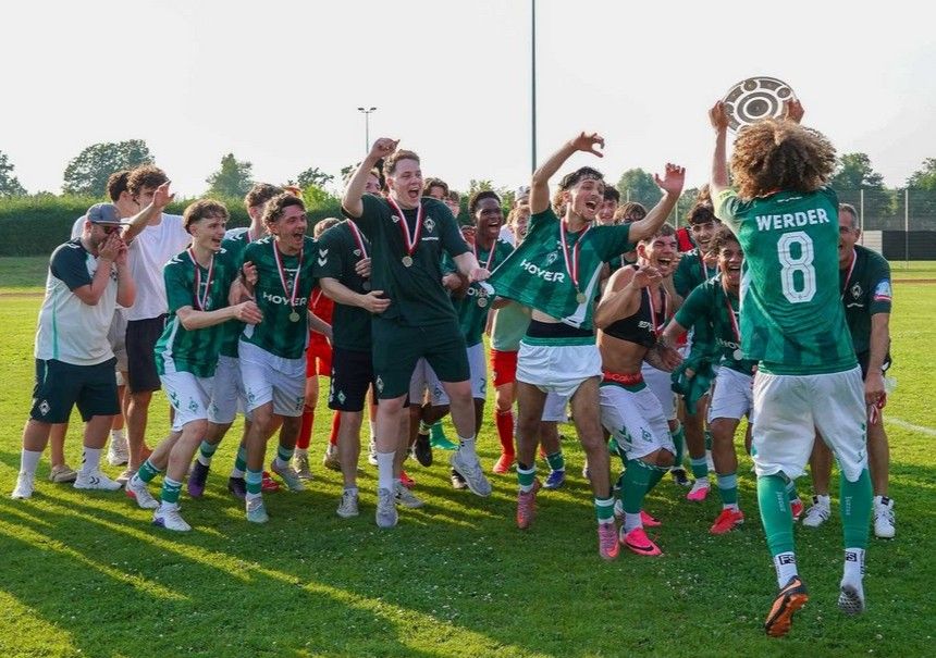 Eine fröhliche Fußballmannschaft in grünen Trikots feiert auf dem Spielfeld; ein Spieler hält einen Pokal in die Höhe, alle tragen Medaillen.