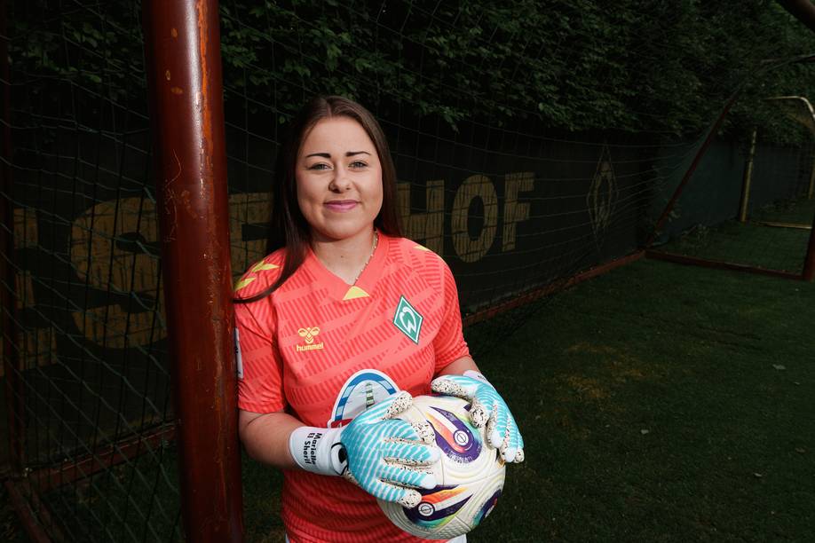 Mariella El Sherif in einem bunten Torwarttrikot, einen Fußball in der Hand, steht neben einem Tor, im Hintergrund ist Grün zu sehen.