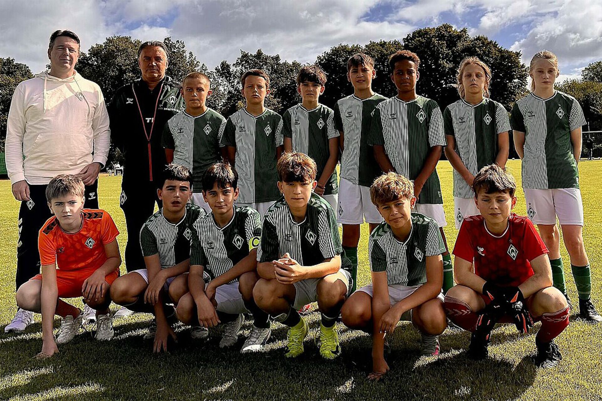 Eine Jugendfußballmannschaft posiert auf einem Spielfeld in grün-weißen Trikots. Hinter ihnen stehen zwei Trainer. Im Hintergrund sind Bäume und ein bewölkter Himmel zu sehen.