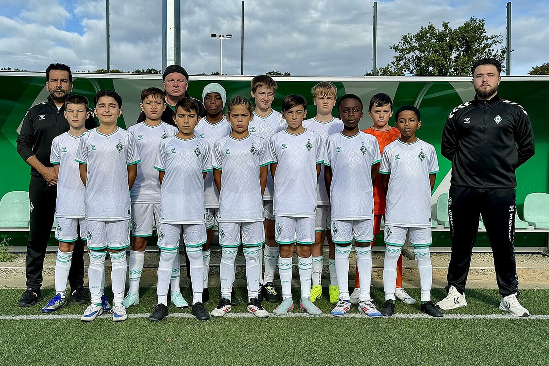 Eine Jugendfußballmannschaft in weißen Trikots posiert für ein Gruppenfoto auf einem Spielfeld, während die Trainer zu beiden Seiten stehen.