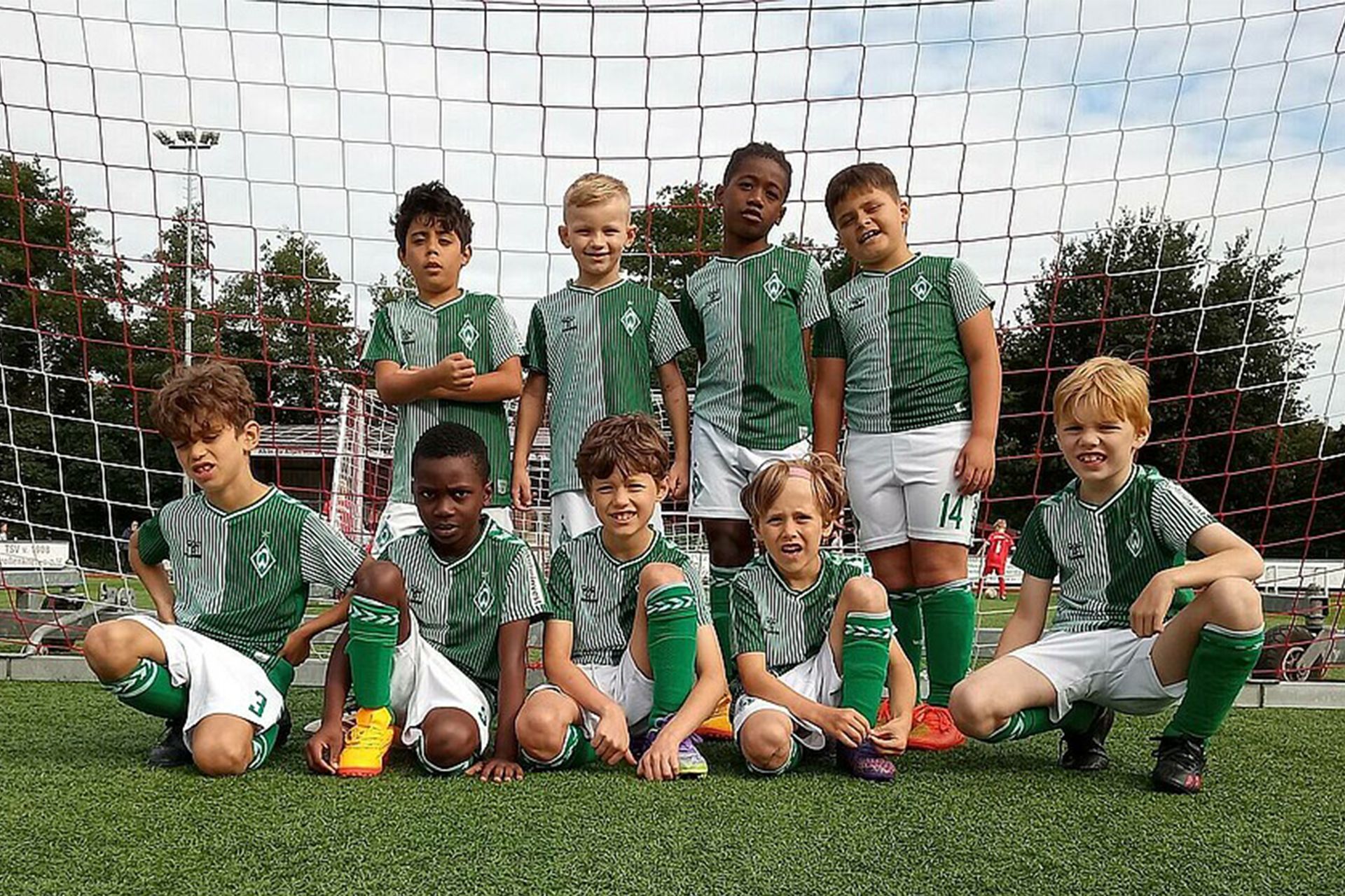 Eine Jugendfußballmannschaft posiert vor einem Tor in grün-weißen Trikots auf einem Rasenfeld mit Bäumen und bewölktem Himmel im Hintergrund.