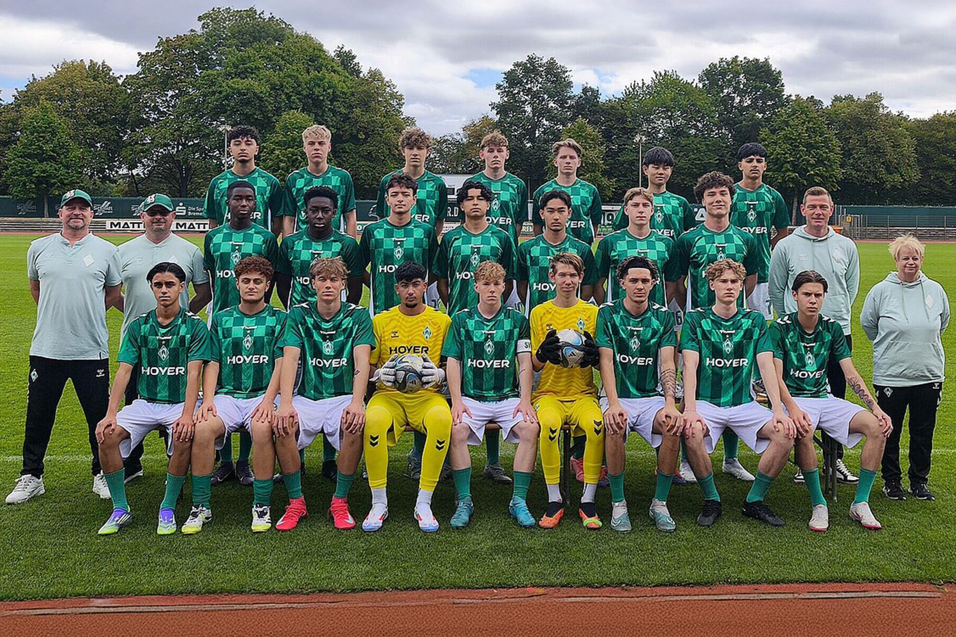 Eine Fußballmannschaft in grün-weißen Trikots posiert mit ihren Trainern auf einem Spielfeld. Zwei Spieler in gelber Torwartkleidung sitzen davor. Im Hintergrund sind Bäume zu sehen.