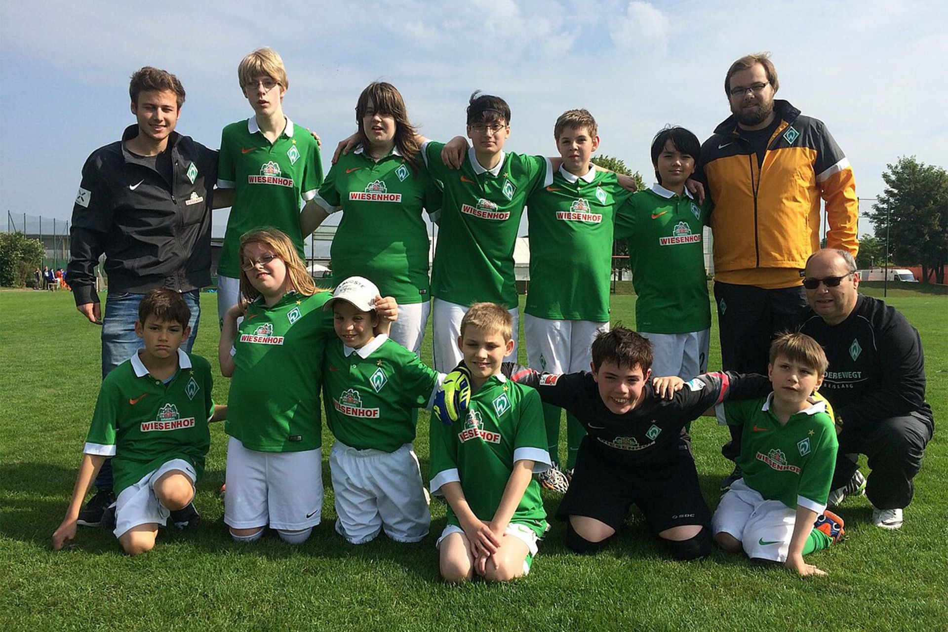 Eine Jugendfußballmannschaft in grünen Trikots posiert auf einem Rasenfeld mit drei Trainern unter einem teilweise bewölkten Himmel.