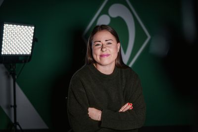 Mariella EL Sherif steht in einem dunklen Pullover und mit verschränkten Armen da und lächelt selbstbewusst. Sie steht vor einem grünen Hintergrund, neben ihr befindet sich eine beleuchtete Tafel.