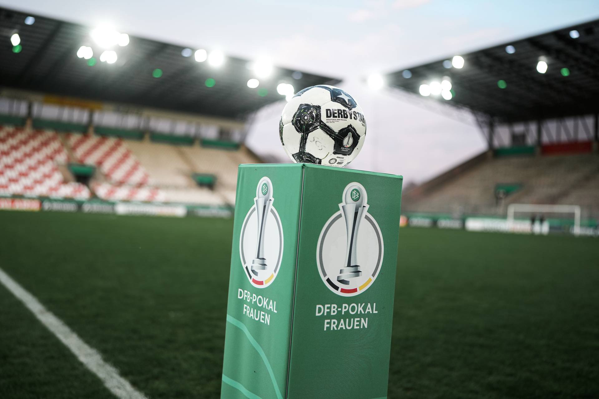 Ein Fußball auf einem grünen Sockel mit dem DFB-Pokal-Frauen-Logo in einem leeren Stadion, beleuchtet von Deckenleuchten.