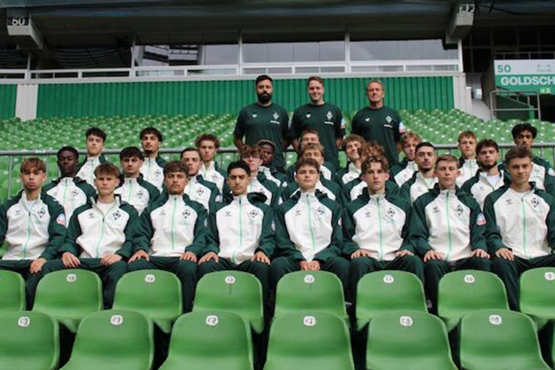 Eine Sportmannschaft posiert für ein Gruppenfoto in einem Stadion. Spieler in weißen und grünen Trainingsanzügen sitzen in Reihen, hinter ihnen stehen die Trainer.