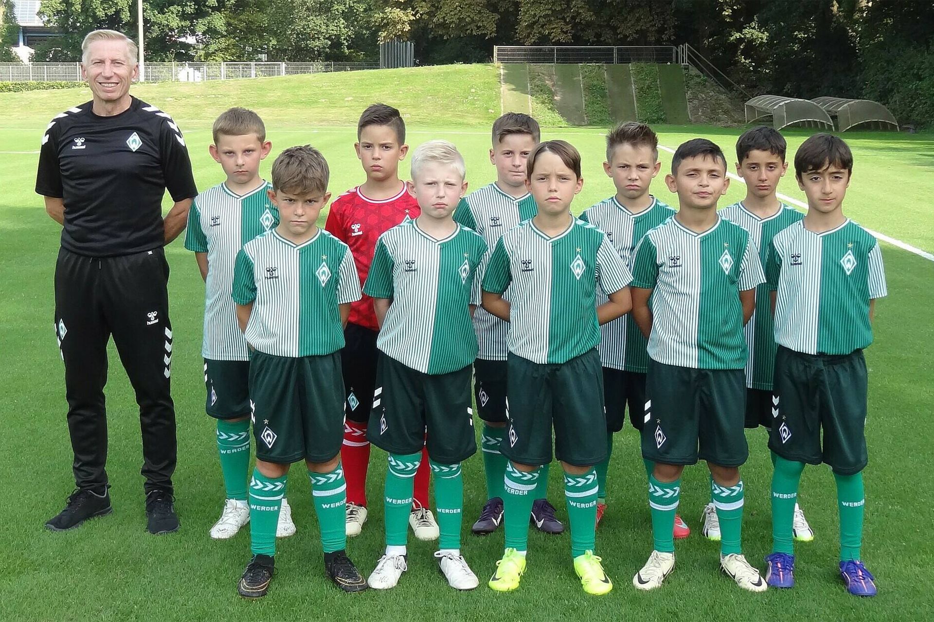 Eine Jugendfußballmannschaft posiert auf einem Spielfeld. Der Trainer steht links, und die Spieler tragen grün-weiße Trikots mit passenden Socken.