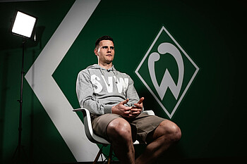 null Michael Zetterer sitzt auf einem Suhl vor einer grünen Wand. Auf der Wand ist die Werder-Raute zu sehen. Hinter ihm steht eine Lampe. Er ist von unten fotografiert. Er trägt eine beige Shorts und einen grauen Pullover.