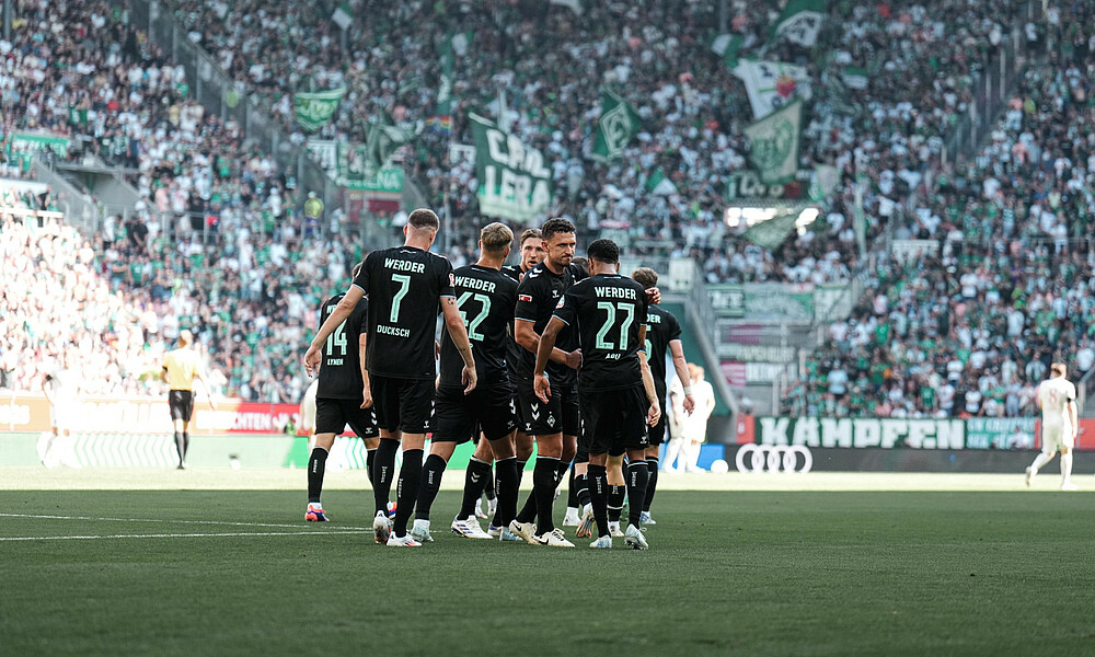 null Die Spieler stehen in einer Traube zusammen. Sie tragen die schwarzen Away-Trikots. Im Hintergrund sieht man den Werder-Block. Es werden viele Fahnen geschwenkt.