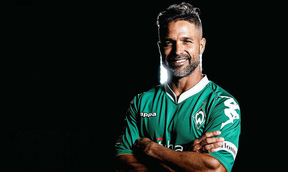 null Diego trägt ein grünes Trikot. Er grinst. Der Hintergrund ist schwarz, hinter ihm ist eine Lampe.