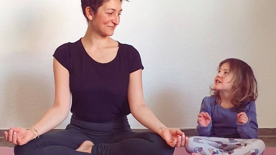Frau und junges Mädchen sitzen mit gekreuzten Beinen auf einer Yogamatte und lächeln sich in einer friedlichen Umgebung an.