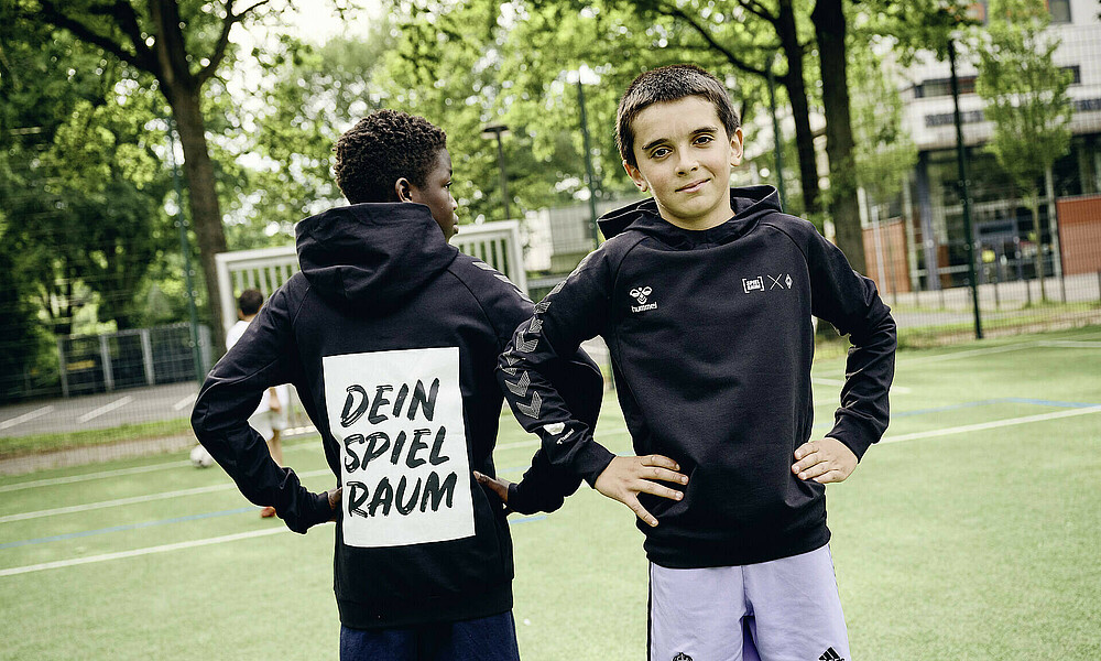 Auf dem Bild sind Jungs im Alter von ungefähr 14 Jahren zu sehen, die auf einem Bolzplatz in SPIELRAUM-Merch stehen.