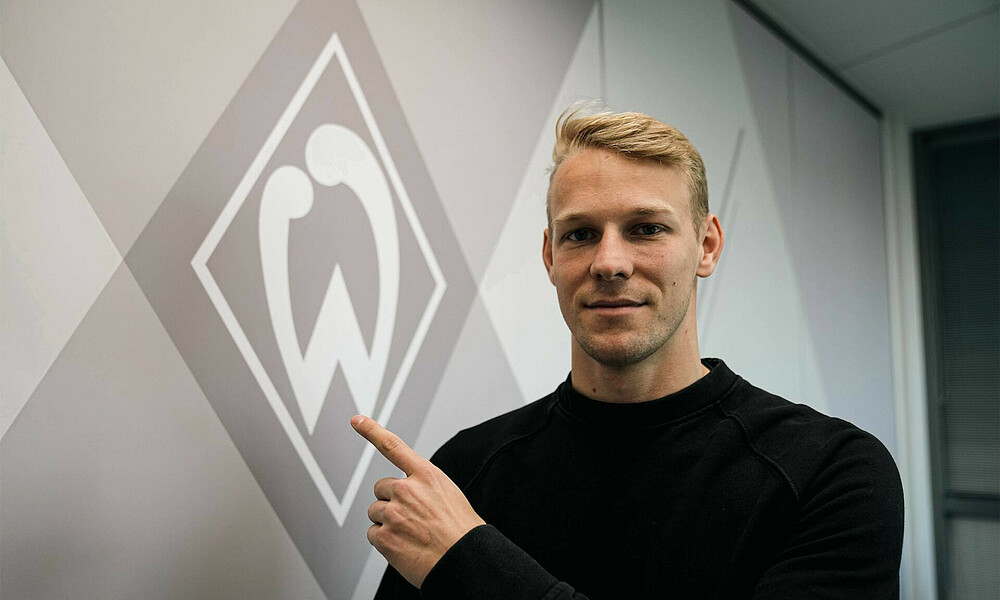 Grüll deutet vor einer Wand auf eine Werder-Raute. 