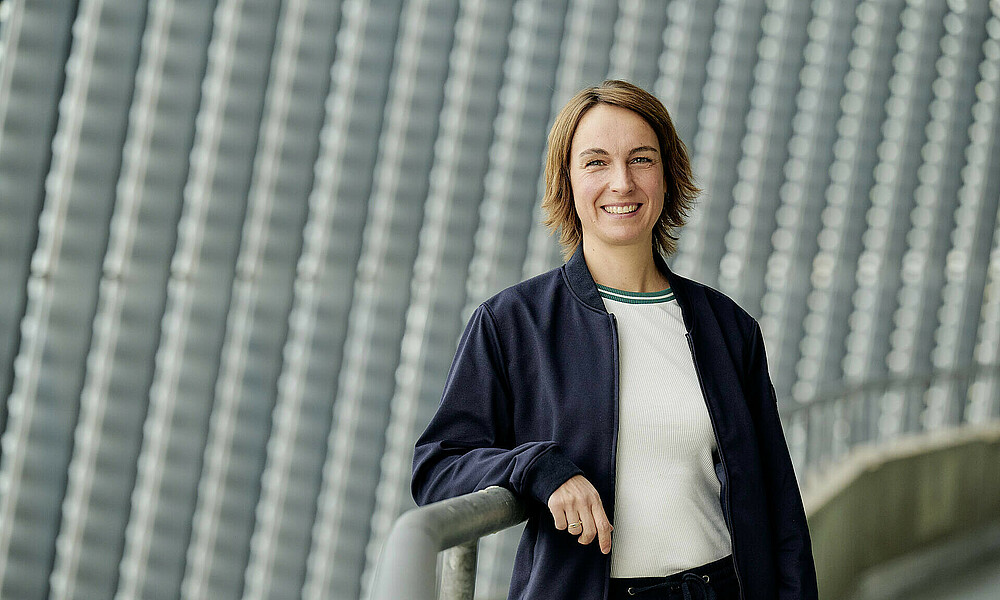 Anne-Kathrin Laufmann lehnt an einem Geländer, im Hintergrund ist die Photoviltaik-Anlage des wohninvest WESERSTADIONs zu sehen.