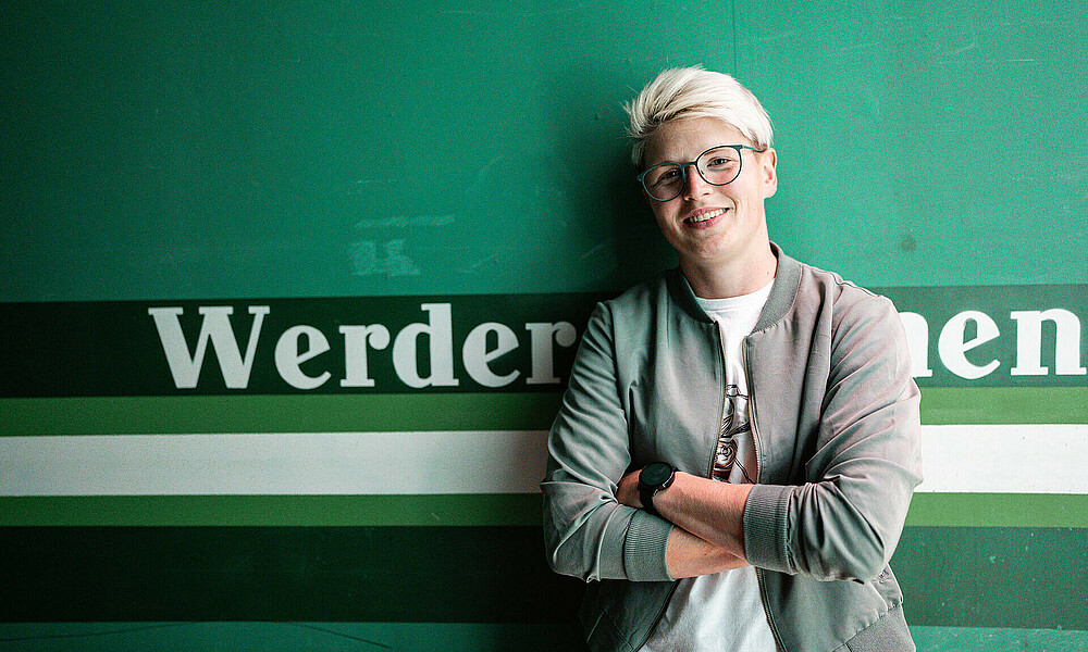 Vanessa Fischer steht mit verschränkten Armen vor einer grünen Wand, auf der 