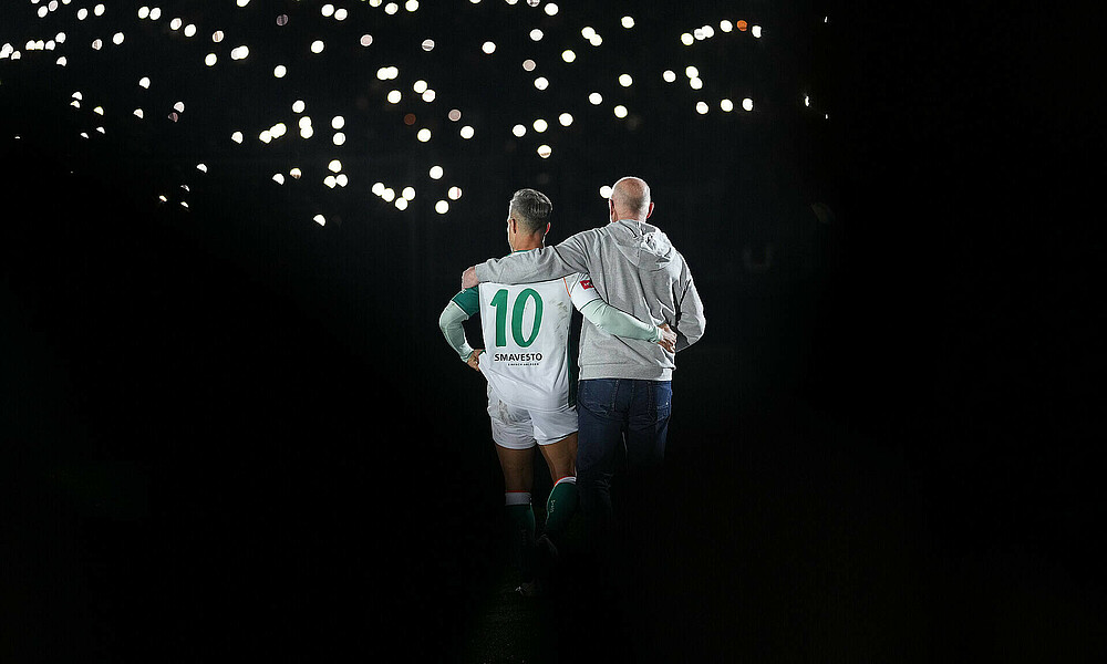 Diego und Thomas Schaaf sind von hinten zu sehen. Der Hintergrund ist dunkel, es leuchten Taschenlampen.