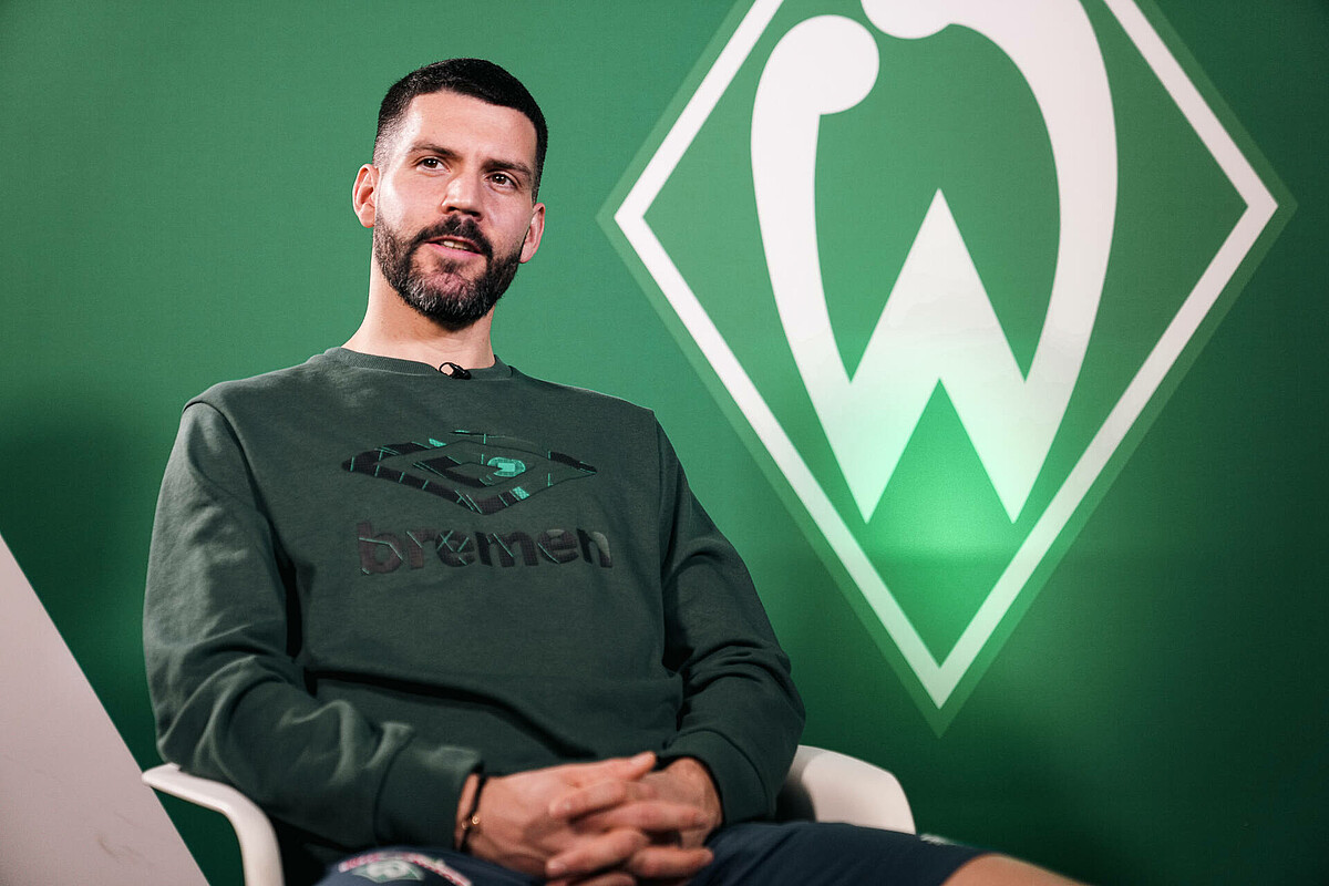 null Anthony Jung sitzt auf einem Stuhl für das Interview - im Hintergrund die Werder-Raute.