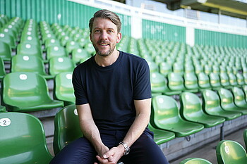 Peter Niemeyer sitzt auf einer grünen Sitzschale auf der Tribüne im Stadion, seine Hände sind gefaltet. Er trägt ein schwarzes T-Shirt. 