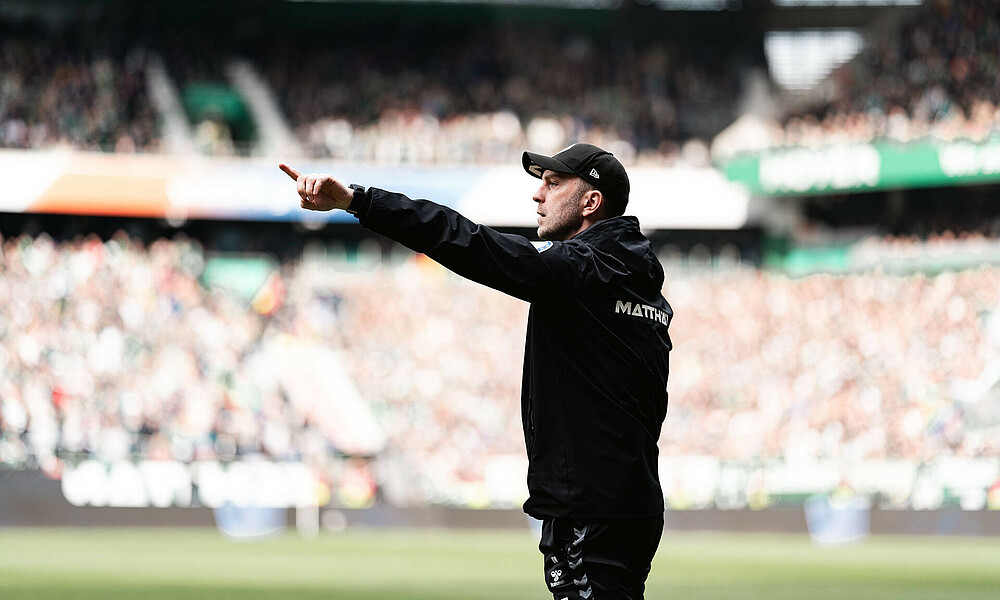 null Ole Werner zeigt mit dem Finger in die Ferne. er steht im Stadion und hat eine schwarze Kappe auf.