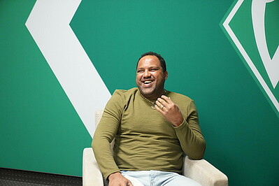 Ailton sitzt im Sessel und lacht. Im Hintergrund die Werder-Raute.