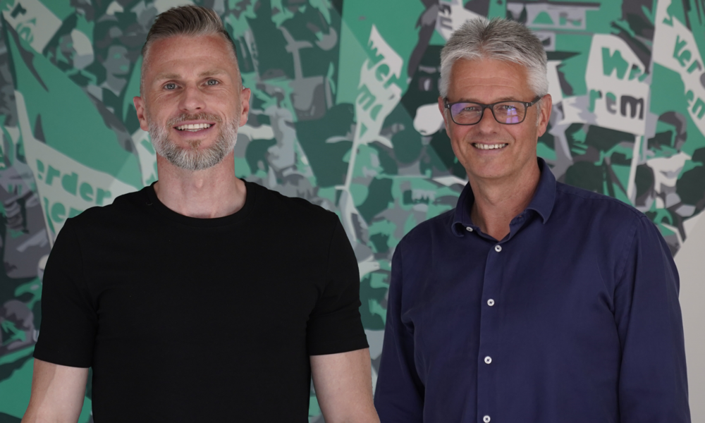 null Daniel Flottmann (li.) und Ralf Heskamp (re.) stehend lächeld vor einer Graffiti-Wand. Auf der Wand sind grün-weiße Werder-Fahnen zu sehen.