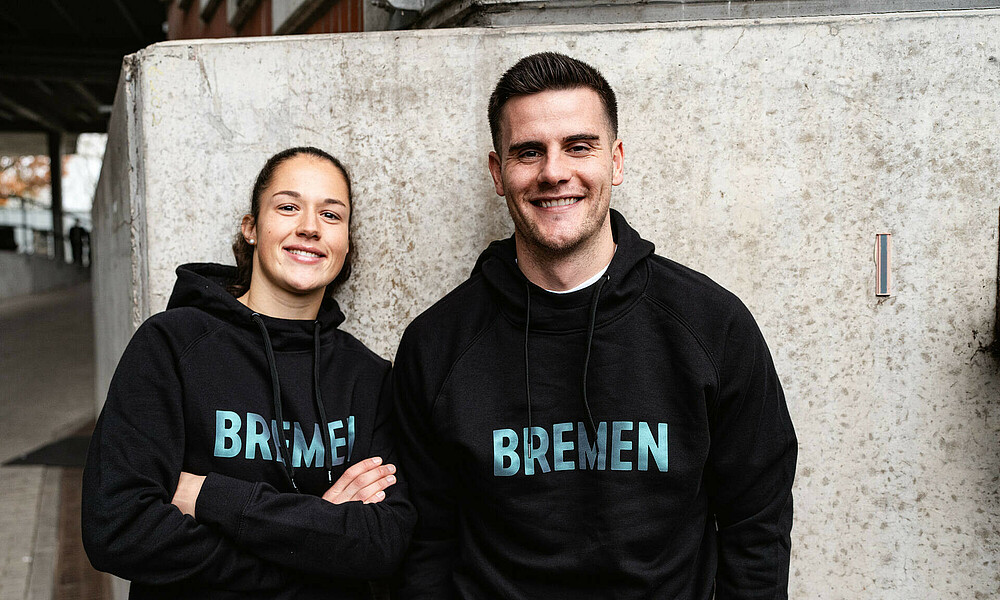 Livia Peng und Michael Zetterer vor einer grauen Wand im Weserstadion im schwarzen Hoody
