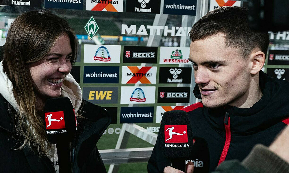 Juliane und Florian Wirtz während eines Interviews vor einer Sponsorenwand. Sie haben beide Mikrofone mit dem Bundesliga-Logo in der Hand.