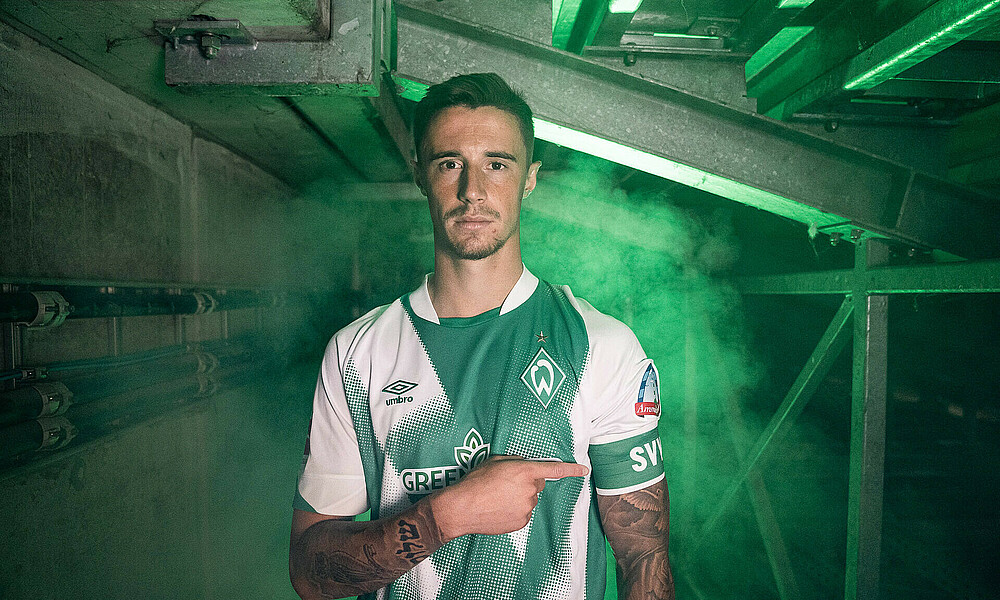 Marco Friedl im neuen HOME-Trikot des SVW. Er trägt die Kapitänsbinde und zeigt mit der rechten Hand darauf.