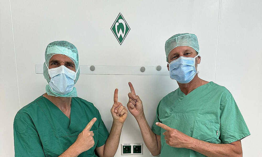 Maximilian Massalme steht mit einem Kollegen in seinem Operationssaal und zeigt auf ein Werder-Logo an der Wand.