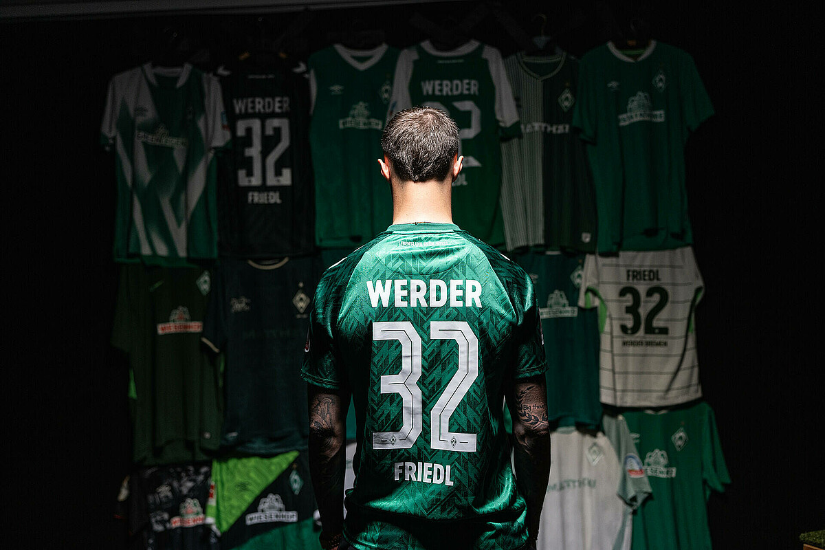 null Marco Friedl ist von hinten zu sehen. Er trägt das grüne Heim-Trikot. Er schaut auf die Wand mit den zahlreichen Trikots.