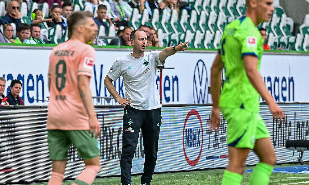 null Ole Werner an der Seitenlinie mit gestrecktem Arm, im Vordergrund unscharf Mitchell Weiser und ein Wolfsburger Spieler