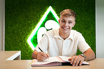 Keke Topp lächelt in die Kamera. Vor ihm liegt eine Mappe. Er hat einen Stift in der Hand. Er trägt ein helles Oberteil und sitzt vor einer grünen Wand.