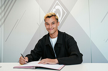 Mio Backhaus unterzeichnet seinen Vertrag und schaut in die Kamera