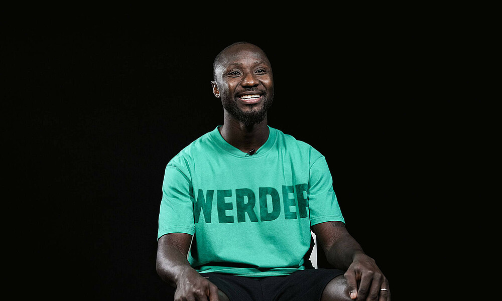 Naby Keïta sitzt in grünem Tshirt vor schwarzem Hintergrund. Er hat die Hände auf die Knie gelegt.