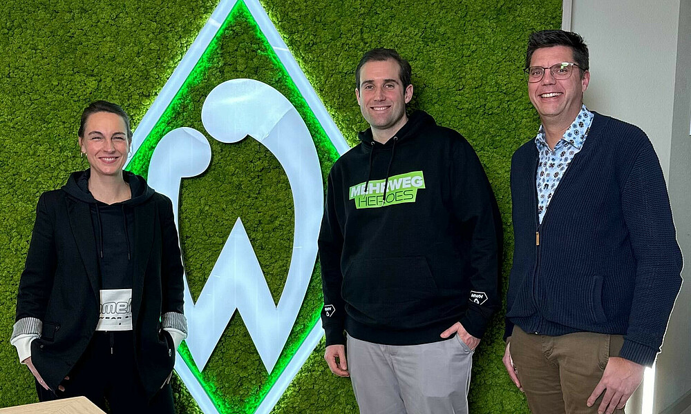Anne-Katrin Laufmann steht gemeinsam mit dem Podcast Hosts vor einem großen Werder Logo