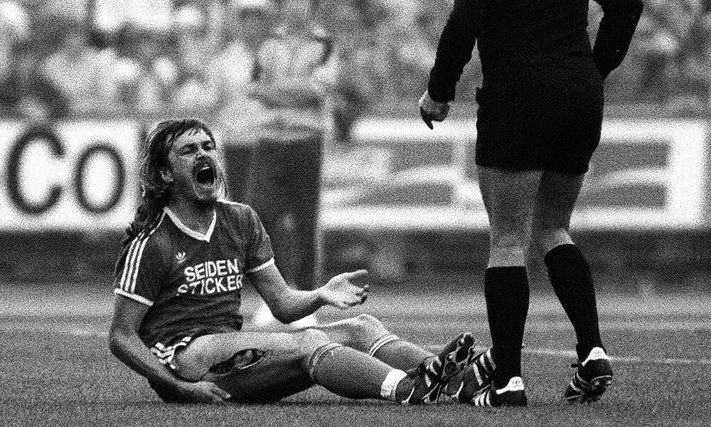 null Ewald Lienen sitzt auf dem Boden, sein Oberschenkel ist offen.