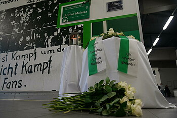 Unter der Gedenktafel befinden sich ein Tisch mit einem Kranz, auf dem Boden liegen zahlreiche weiße Rosen.