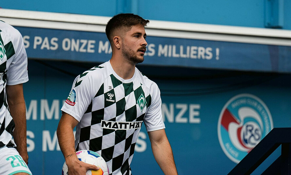 Romano Schmid im grün-weiß-karierten Aufwärmshirt mit einem Ball unter den rechten Arm geklemmt.