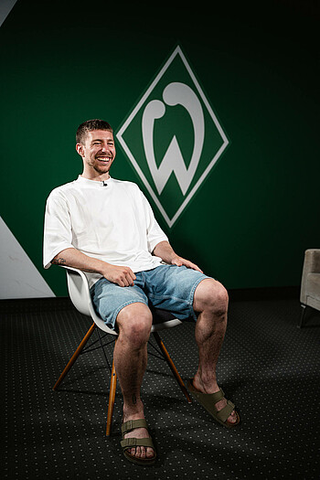 null Mitchell weiser lachend im Interview. Er sitzt auf einem weißen Stuhl. Er trägt ein weißes Tshirt, Jeansshorts und Latschen.