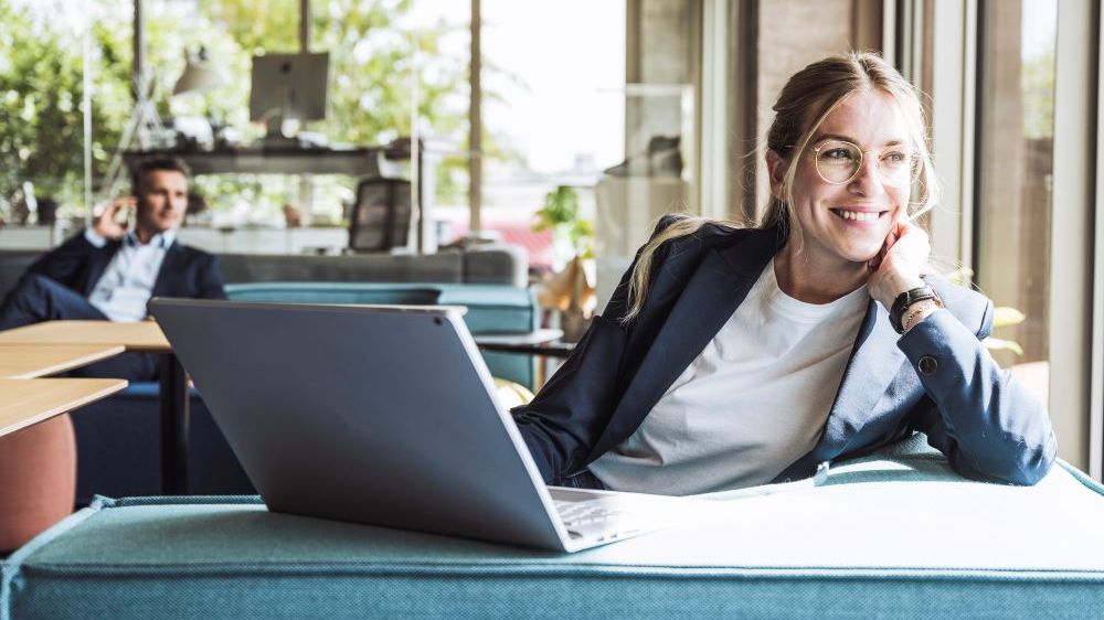 Aktienfonds investieren Blonde Businessfrau mit Brille mit aufgeklapptem Notebook blickt lächelnd aus dem Fenster. Im Hintergrund sieht man einen Kollegen